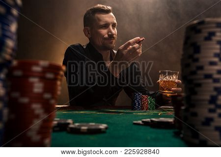 Young Handsome Man Sitting Behind Poker Table With Cards And Chips. In The Foreground Stacks Of Chip