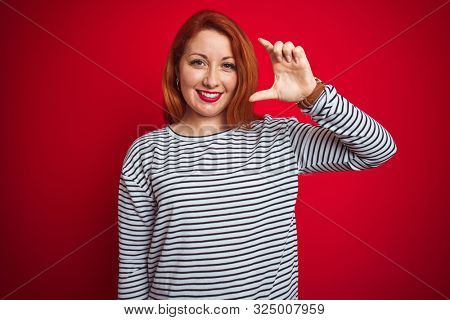 Young redhead woman wearing strapes navy shirt standing over red isolated background smiling and confident gesturing with hand doing small size sign with fingers looking and the camera.