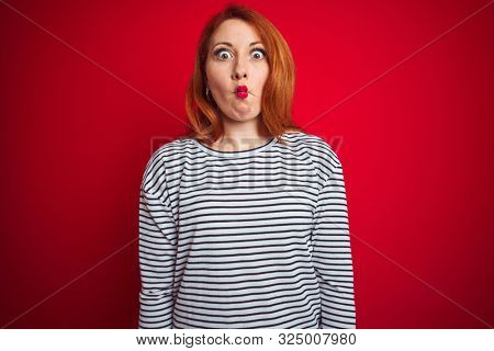 Young redhead woman wearing strapes navy shirt standing over red isolated background making fish face with lips, crazy and comical gesture. Funny expression.