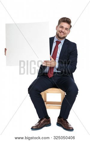 happy young businessman smiling and holdy empty white board, sitting isolated on white background in studio