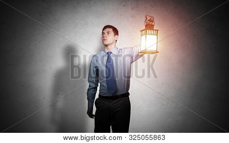 Businessman With Glowing Lantern Looks Suspicious On Background Grunge Wall. Young Corporate Employe