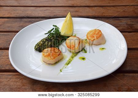 Close Up Portion Of Three Grilled Scallops On White Plate Over Brown Wooden Table, High Angle View