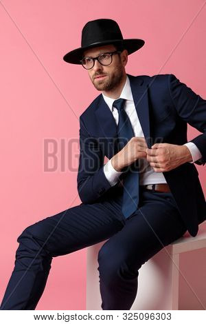mysterious formal business man wearing a navy suit,black hat,glasses is sitting and grabbing something from his inner jacket pocket while looking away serious on pink studio background