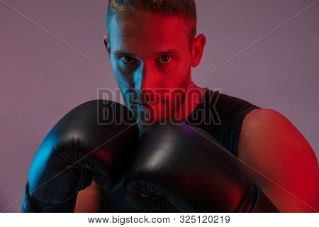 Photo of a concentrated strong young sports man boxer posing isolated over neon lights in gloves make boxing exercises.