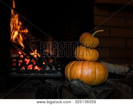 Three Halloween Pumpkins Stand On Top Of Each Other On Birch Firewood Next To A Flaming Fireplace.