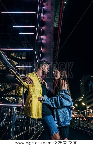 Side View Of Handsome Boyfriend And Attractive Girlfriend In Denim Jacket Hugging In Night City