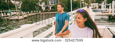 Happy young people couple walking on Florida boardwalk in Marina harbour pier holding hands. Interracial relationship Asian girl, Caucasian man enjoying holiday. Panoramica banner.