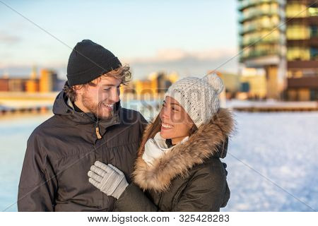 Winter couple walking happy in city wearing cold outerwear snow hat and jackets. Young biracial adults together smiling looking at each other on walk. Asian girlfriend in love with man.