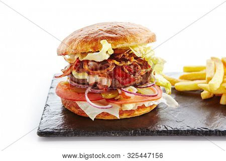 Fresh Beef Burger with Bacon, Fried Onions, Tomato Sauce, Pickled Cucumbers, Green Lettuce, Cheese and French Fries Garnish. Hamburger or Beefburger on Natural Black Stone Plate Isolated on White