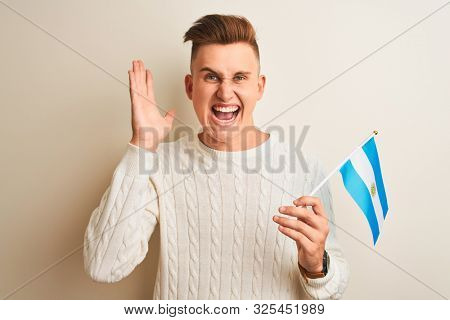 Young handsome man holding Argentinian Argentina flag over isolated white background very happy and excited, winner expression celebrating victory screaming with big smile and raised hands