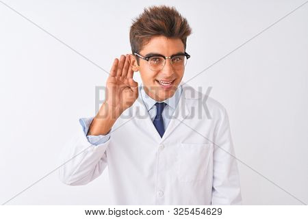 Young handsome sciencist man wearing glasses and coat over isolated white background smiling with hand over ear listening an hearing to rumor or gossip. Deafness concept.