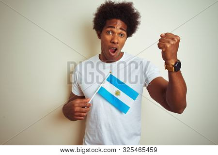 Afro american tourist man holding Argentinian Argentina flag over isolated white background annoyed and frustrated shouting with anger, crazy and yelling with raised hand, anger concept