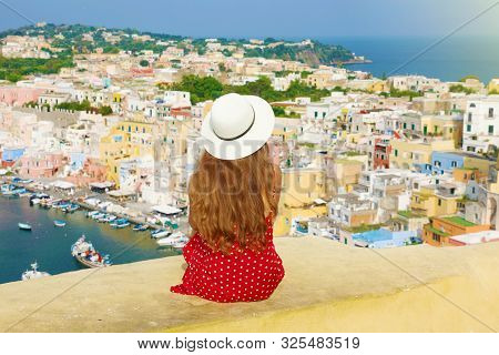 Travels In Italy. Back View Beautiful Young Woman With Hat Sitting On Wall Looking At Stunning Panor