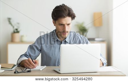 Budget Planning. Serious Man Working On Laptop And Taking Notes Indoor.
