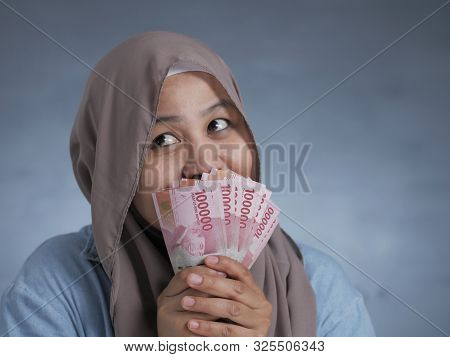 Portrait Of Indonesian Muslim Woman Holding Rupiah Money, Smiling And Thinking Gesture