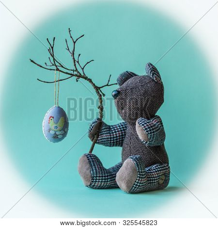 Toy Teddy Bear Sitting On The  Turquoise Background With A Rough Branch And  Easter Decoreted Egg.