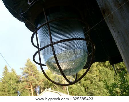 Close Up Of A Street Glass Lantern In A Metal Protective Vandal Proof Cage. In The Background Are Vi