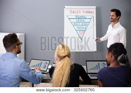 Young Smiling Businessman Explaining Sales Funnel Diagram To His Colleagues In Meeting