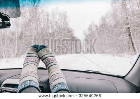 Woman feet in warm woolen socks on car dashboard over snow view. Having weekend trip in winter forest. Freedom travel concept.
