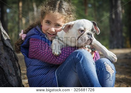 In The Autumn Forest, A Little Girl Holds A French Bulldog In Her Arms. She Is Happy, Hugs Him.