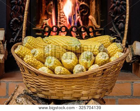 A Wicker Basket To The Top Filled With Ears Of Ripe Corn. In The Background Firewood Burning In The