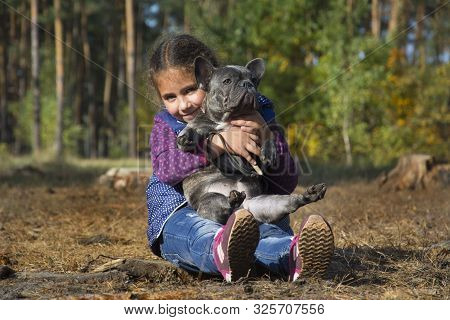 In The Autumn Forest, A Little Girl Holds A French Bulldog In Her Arms. She Is Happy, Hugs Him.