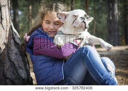 In The Autumn Forest, A Little Girl Holds A French Bulldog In Her Arms. She Is Happy, Hugs Him.they 