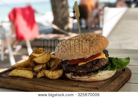 Grilled Burger On Wooden Board With Salad And Potato Chips
 With A Beach In The Background Out Of Fo
