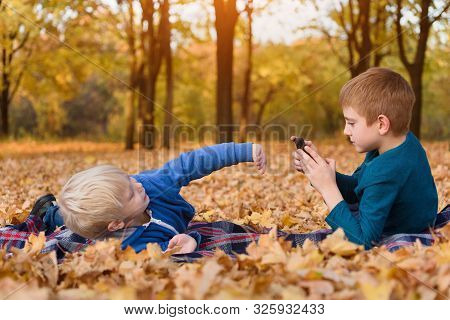Two Little Brothers Take Pictures Of Each Other, Lying In Yellow Autumn Leaves. Fall Day
