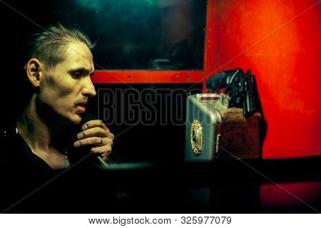A Man Of Criminal Appearance In A Payphone Booth At Night. Talking On The Phone With A Revolver And