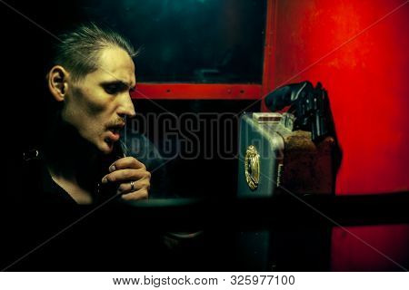 A Man Of Criminal Appearance In A Payphone Booth At Night. Talking On The Phone With A Revolver And