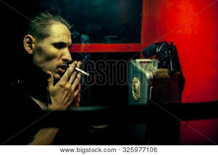 A Man Of Criminal Appearance In A Payphone Booth At Night. Talking On The Phone With A Revolver And