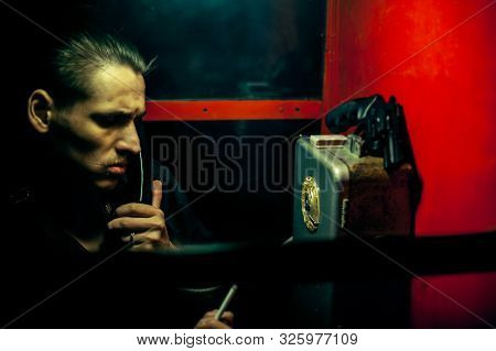 A Man Of Criminal Appearance In A Payphone Booth At Night. Talking On The Phone With A Revolver And