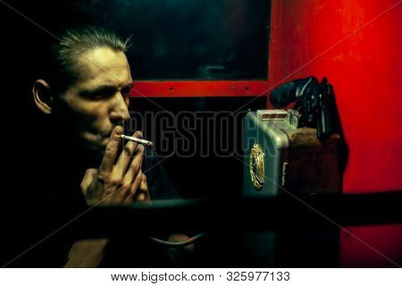 A Man Of Criminal Appearance In A Payphone Booth At Night. Talking On The Phone With A Revolver And