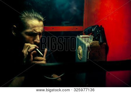 A Man Of Criminal Appearance In A Payphone Booth At Night. Talking On The Phone With A Revolver And