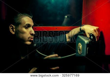 A Man Of Criminal Appearance In A Payphone Booth At Night. Talking On The Phone With A Revolver And