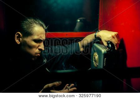 A Man Of Criminal Appearance In A Payphone Booth At Night. Talking On The Phone With A Revolver And