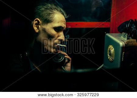 A Man Of Criminal Appearance In A Payphone Booth At Night. Talking On The Phone With A Revolver And