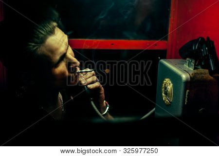 A Man Of Criminal Appearance In A Payphone Booth At Night. Talking On The Phone With A Revolver And