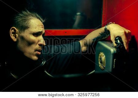 A Man Of Criminal Appearance In A Payphone Booth At Night. Talking On The Phone With A Revolver And