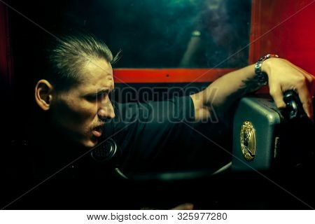 A Man Of Criminal Appearance In A Payphone Booth At Night. Talking On The Phone With A Revolver And