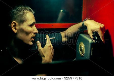 A Man Of Criminal Appearance In A Payphone Booth At Night. Talking On The Phone With A Revolver And