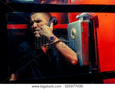 A Man Of Criminal Appearance In A Payphone Booth At Night. Talking On The Phone With A Revolver And