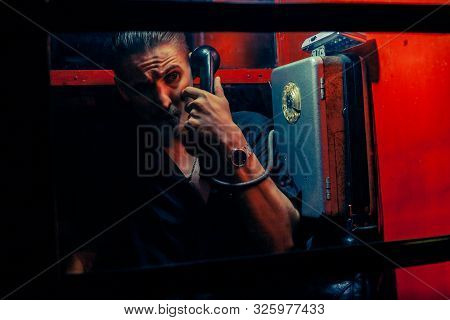 A Man Of Criminal Appearance In A Payphone Booth At Night. Talking On The Phone With A Revolver And