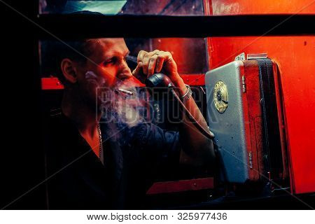 A Man Of Criminal Appearance In A Payphone Booth At Night. Talking On The Phone With A Revolver And