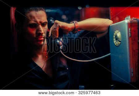 A Man Of Criminal Appearance In A Payphone Booth At Night. Talking On The Phone With A Revolver And