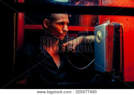 A Man Of Criminal Appearance In A Payphone Booth At Night. Talking On The Phone With A Revolver And