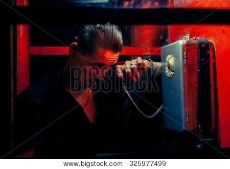 A Man Of Criminal Appearance In A Payphone Booth At Night. Talking On The Phone With A Revolver And