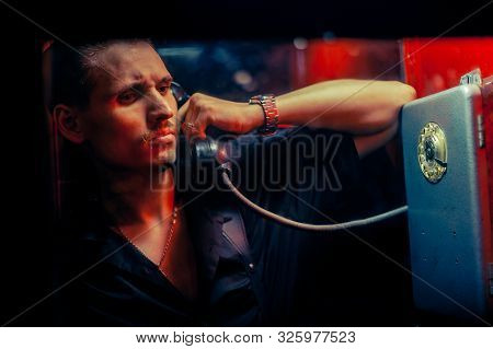 A Man Of Criminal Appearance In A Payphone Booth At Night. Talking On The Phone With A Revolver And