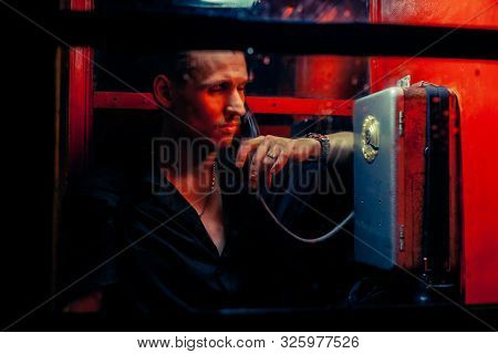 A Man Of Criminal Appearance In A Payphone Booth At Night. Talking On The Phone With A Revolver And